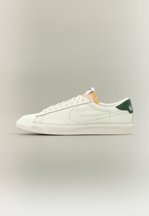 Valkoinen matala tennari, jossa on rei’itetty Nike-swoosh, valkoiset nauhat, vihreä kantapään lisäosa valkoisella Nike-tekstillä ja näkyvissä oleva oranssi kielen vaahto.