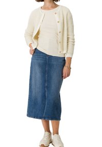 Hellbeige, flauschige Strickjacke mit goldenen Knöpfen, kombiniert mit einem gerippten weißen Oberteil und einem blauen Denim-Midirock. Weiße Sneaker runden das Outfit ab.