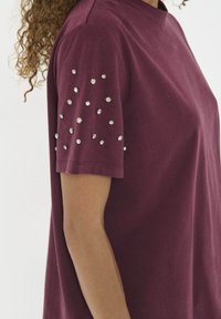 Ženska oblečena v kratkosleeve maroon srajco z razpršenimi okroglimi srebrnimi žeblji na rokavu, prikazana od rame do sredine trupa.