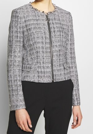 Femme portant une veste zippée texturée noire et blanche avec bordure à franges, associée à un pantalon noir sur un fond uni.