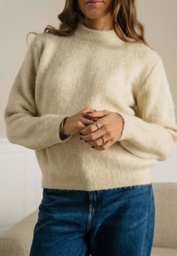 Cremefarvet uldtrøje med høj hals og ribbet kant. Parret med blå denimjeans. Hænderne er foldet, og flere ringe vises.