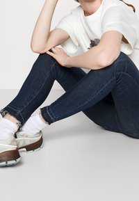 Tunna mörkblå jeans med fransade fållar, i kombination med beige och bruna sneakers. Modellen sitter med benen korsade, klädd i en vit t-shirt med grafik.