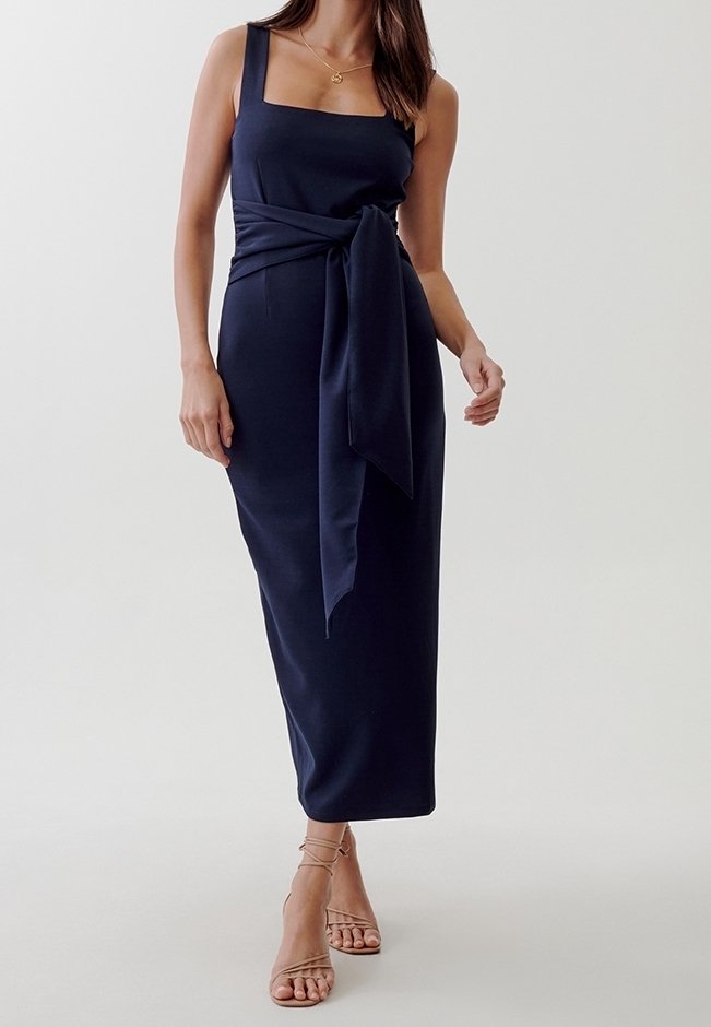 Femme portant une robe midi sans manches bleu marine avec une large ceinture à la taille et un décolleté carré, associée à des talons nus à brides sur un fond uni.