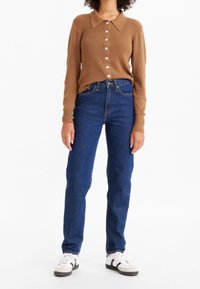 Kvinna som bär en brun ribbad knappad tröja, blå högmidjade jeans och vita sneakers med svarta ränder, stående mot en vit bakgrund.