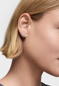 Pendiente de clip en forma de flor de plata con un acabado suave, situado en el lóbulo de la oreja, que complementa un peinado corto y rubio.
