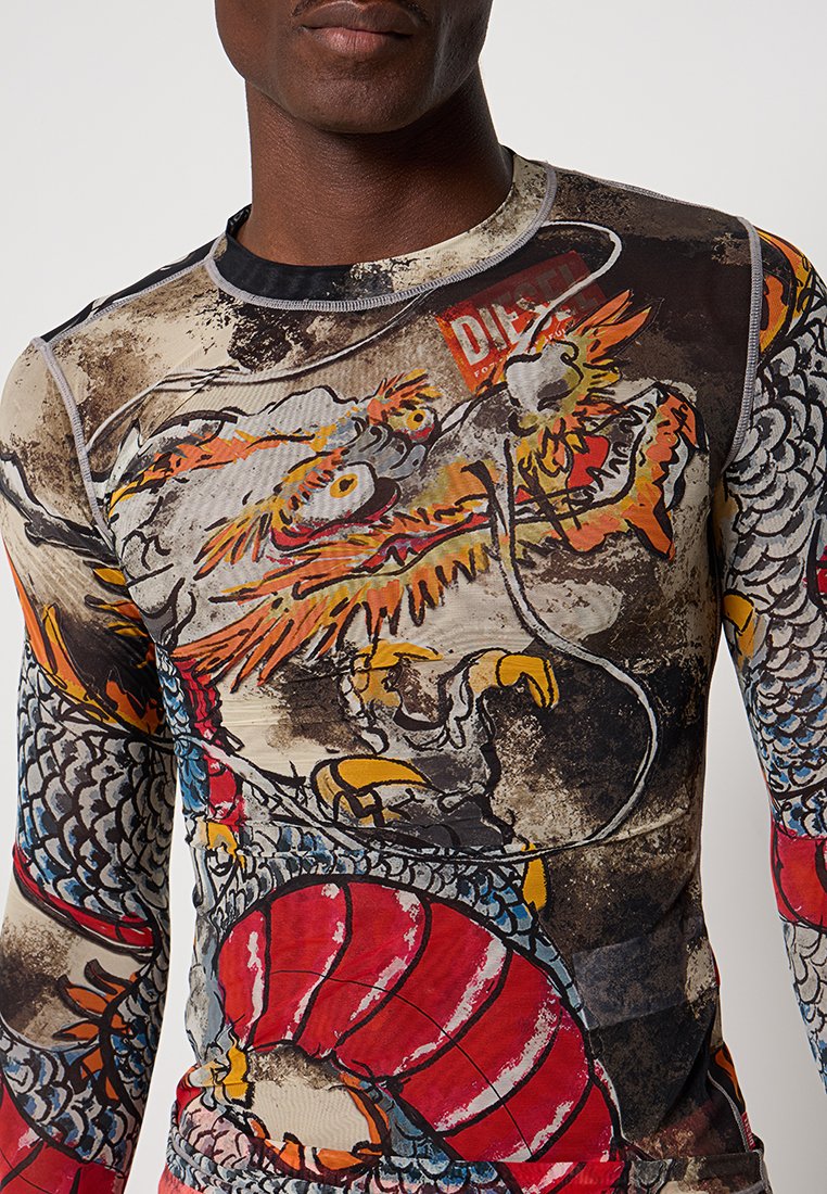 Ein Mann trägt ein Langarmshirt mit bunten Drachenmotiven in Orange, Rot, Gelb und Blau auf einem strukturierten beigen und schwarzen Hintergrund.