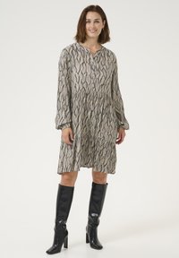 Beige pitkähihaine mekko mustalla aalto kuvioinnilla, napitettava etuosa, kerätty vyötärö, yhdistetty polveen ulottuviin mustiin saappaisiin. Suuri kangas.