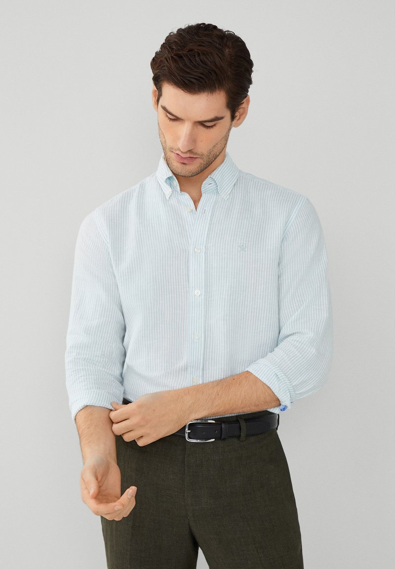 Camisa de botones a rayas azul claro con cuello, mangas remangadas y un pequeño logo en el pecho, combinada con pantalones oscuros y un cinturón negro.