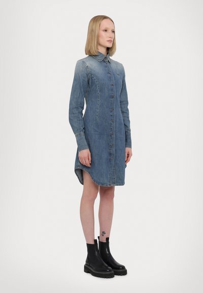 Denim kleita, zila, garas piedurknes, ar pogām priekšā, nedaudz izliekta apakšmala. Stilizēta ar melniem potīšu zābakiem un redzamu tetovējumu uz kājas.