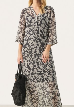 Femme portant une robe midi noire et blanche à fleurs en tissu transparent avec des manches trois-quarts, tenant un sac fourre-tout noir tissé.