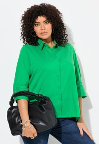 Blusa verde con cuello y mangas de tres cuartos; textura suave. Bolso negro con un asa anudada. Jeans de mezclilla azul.