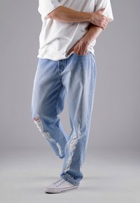 Person, der hellblaue, zerrissene Jeans, ein weißes T-Shirt und weiße Sneaker trägt, steht mit einer Hand in der Tasche und der anderen auf dem Arm.