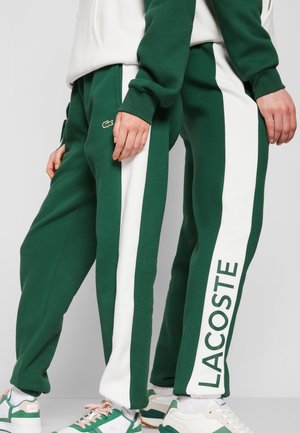 Dwie osoby ubrane w zielone i białe dresy Lacoste z wyraźnymi bocznymi paskami oraz sneakersy z logo Lacoste stoją obok siebie.