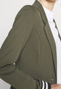 Blazer verde oliva con un design aderente, caratterizzato da una texture liscia e dettagli di cucitura sottili. Camicia bianca sotto con colletto a volant.