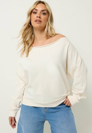Lyshåret kvinde iført løs hvid off-shoulder sweater og blå jeans, poserer med den ene hånd i lommen mod en ensfarvet baggrund.