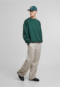 Felpa verde, pantaloni beige a gamba larga, scarpe nere robuste e un cappellino verde con logo bianco. I tessuti sembrano morbidi e texture.