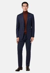 Traje azul marino con un diseño texturizado, combinado con un suéter de cuello alto marrón. Incluye un pañuelo de bolsillo y zapatos marrones pulidos. Ajuste a medida.