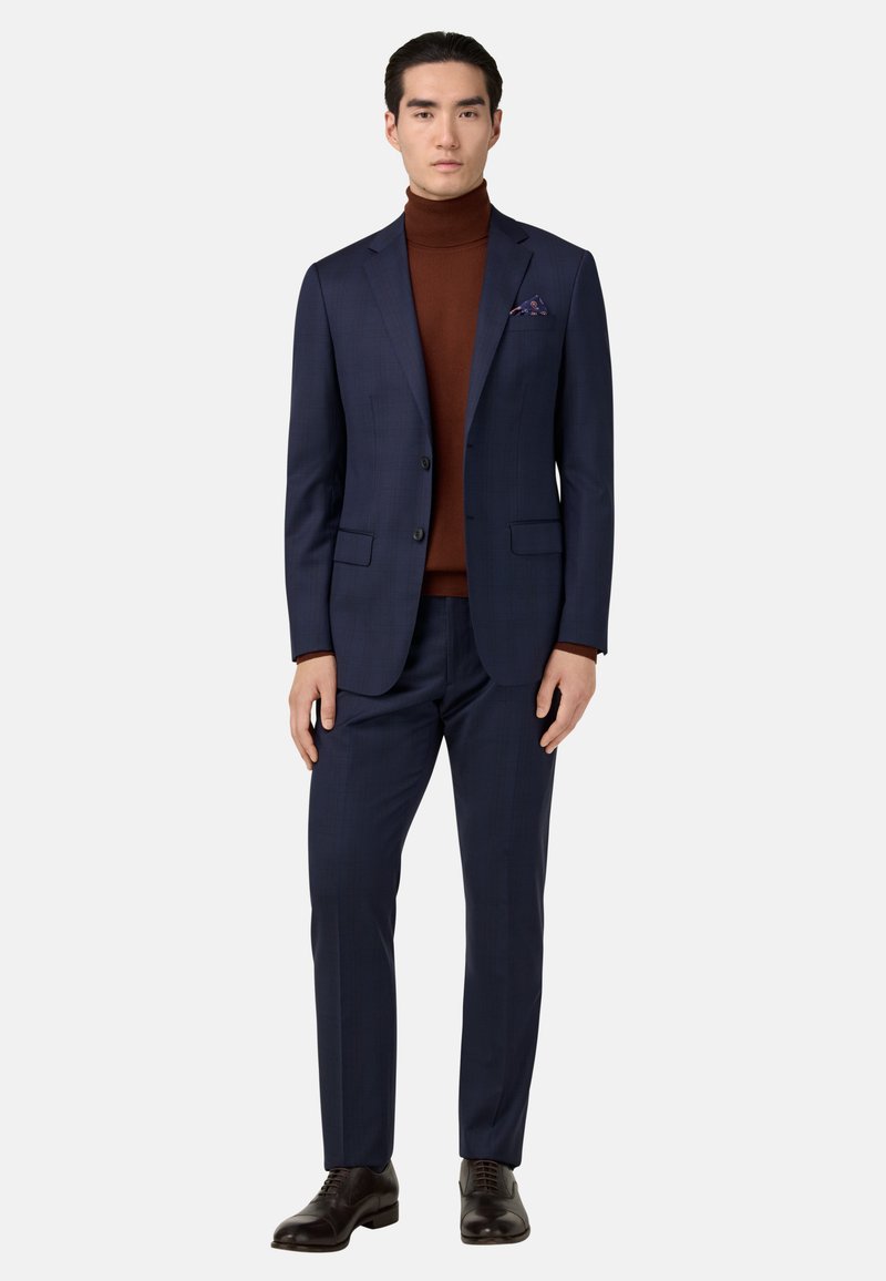 Traje azul marino con un diseño texturizado, combinado con un suéter de cuello alto marrón. Incluye un pañuelo de bolsillo y zapatos marrones pulidos. Ajuste a medida.