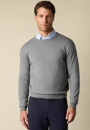 Grauer Pullover mit rundem Ausschnitt und langen Ärmeln, aus einem weichen Stoff. Über einem hellblauen Hemd mit Kragen getragen, mit gerippten Bündchen und Saum.