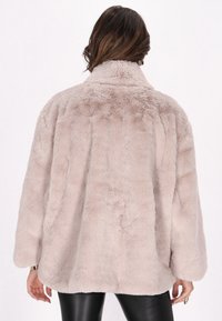 Jachetă din blană artificială într-o nuanță moale de bej, având un guler înalt, o croială lejeră și mâneci lungi. Partea din spate prezintă un design texturat, pufoș.