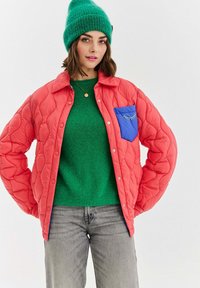 Žena s zelenou pletenou čepicí a svetrem, korálově prošívanou bundou s modrým kapsou a šedými džínami, stojící s rukama v bok.