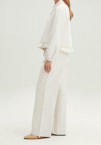 Mujer vestida con una blusa de manga larga color blanco roto con detalles de encaje y pantalones de pierna ancha a juego con flecos, combinados con bailarinas beige.