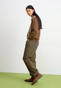 Brown sweater with green accents, green cargo pants, and brown ankle boots. Long straight hair; model poses sideways. Simple background.