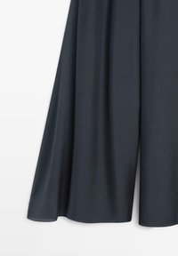 Donkergrijze broek met wijde pijpen, van een losse, vloeiende stof en een gladde textuur, afgebeeld tegen een effen lichte achtergrond.