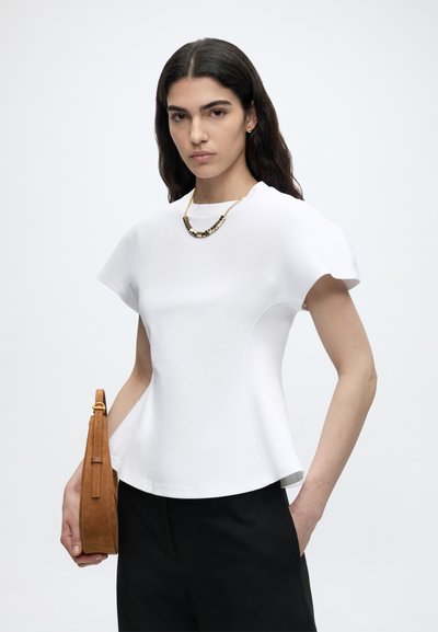 Γυναίκα με μακριά σκουρόχρωμα μαλλιά, που φοράει λευκό εφαρμοστό τοπ, μαύρο παντελόνι, χρυσό κολιέ και κρατά καφέ τσάντα σε λιτό ανοιχτό φόντο.