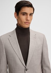 Hellgrauer Blazer mit subtiler Textur, Ein-Knopf-Verschluss und Reverskragen über einem dunkelbraunen Rollkragenpullover.