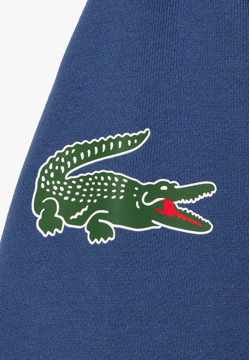 Grünes Krokodil-Logo mit geöffnetem roten Mund auf strukturiertem blauen Stoffhintergrund.