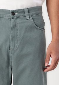 Pantaloni din denim verde deschis cu talie înaltă, buzunare laterale și cusături contrastante. Materialul are o ușoară textură și un croi lejer.