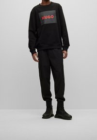 Černá mikina s nápisem "HUGO" červeně, doplněná černými cargo kalhotami s bočními kapsami a černými sportovními botami. Celkově hladká textura.