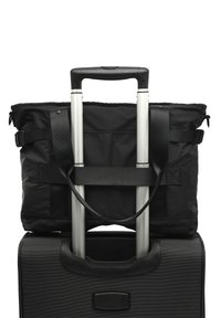 Crni najlon torba s podesivim remenima, osigurana na vrhu crnog kofera s kotačima koji ima teksturiranu površinu i sklopivu ručku.