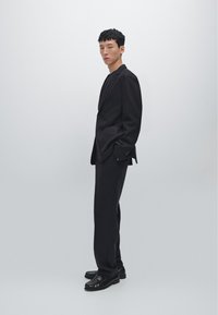 Costume noir sur mesure avec un blazer structuré et un pantalon assorti. Le tissu semble lisse et texturé. Porté avec des mocassins noirs.