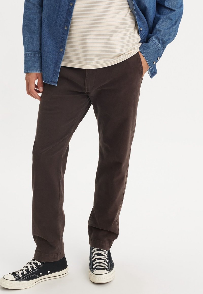 Homme portant un pantalon marron, des baskets noires et blanches, une chemise rayée et une veste en jean bleue, une main dans la poche.