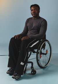 Fekete sportnadrág fehér részletekkel, szürke hosszúujjú póló és fekete sportcipő. Az egyén egy kerekesszékben ül.