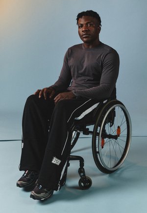 Schwarze Sporthosen mit weißen Akzenten, ein graues Langarmshirt und schwarze Turnschuhe. Die Person sitzt in einem Rollstuhl.
