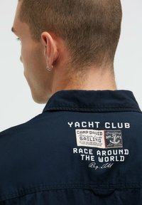 Marineblaues Hemd mit Kragen, bedruckt auf der Rückseite, darunter "Yacht Club" und ein Aufnäher mit nautischen Details.