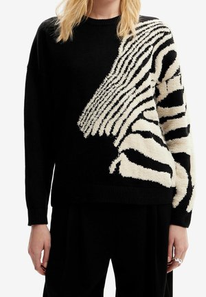 Zwarte truien met crèmekleurig textuur abstract zebra patroon op de rechter schouder en mouw, gedragen met zwarte broek.