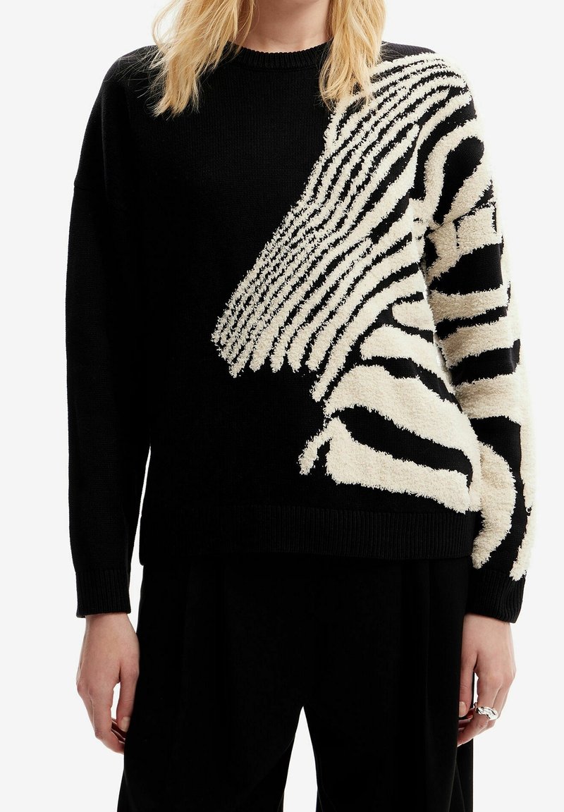 Czarny sweter z kremowym teksturowanym abstrakcyjnym wzorem zebry na prawym ramieniu i rękawie, noszony z czarnymi spodniami.