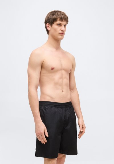 Jeune homme aux cheveux châtains courts portant un short de bain noir, debout devant un fond gris clair uni, regardant légèrement vers la droite.