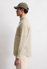 Beige linnen overhemd met knopen en een borstzak, gecombineerd met een groene pet. Het overhemd heeft lange mouwen en een comfortabele pasvorm.