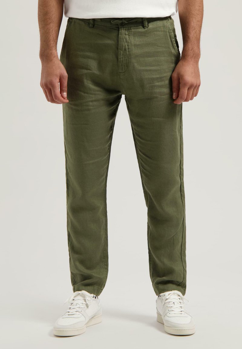 Pantalons en lin léger de couleur vert olive avec une coupe décontractée, dotés d'une taille à cordon et d'accents sur les coutures latérales, associés à des baskets blanches.