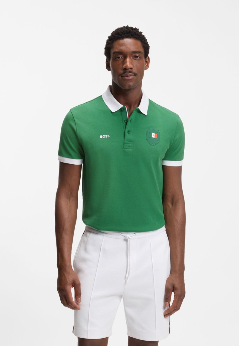 Homme portant un polo vert avec col et poignets blancs, texte « BOSS » et un petit emblème, associé à un short blanc à cordon.