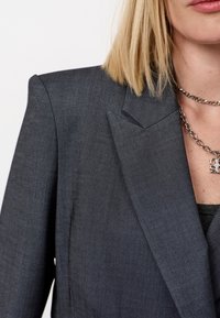 Grijze op maat gemaakte blazer met een gestructureerde revers, een soepele textuur en scherpe schouderlijnen, gecombineerd met een zilveren ketting.