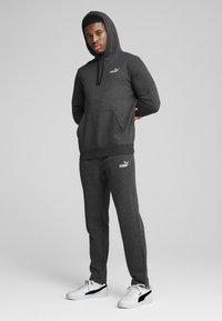 Jonge man in een donkergrijze Puma-hoodie en bijpassende sweatpants met witte Puma-sneakers, staand voor een effen grijze achtergrond.