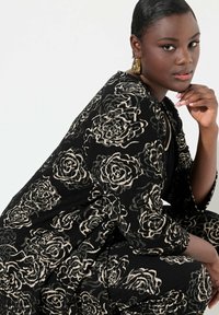 Musta kangaspuku, jossa on teksturoitu kukkakuosisuus kremetoneissa, löysä malli ja kolmannen neljänneksen hihat. Kullansävyiset korvakorut tuovat sävyä.