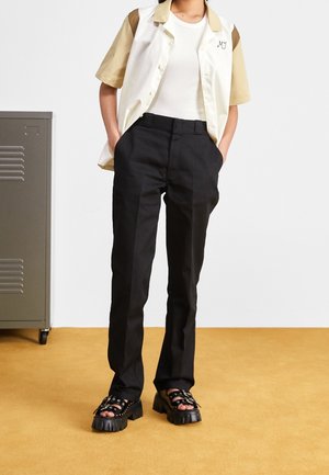 Chemise à manches courtes crème et beige, pantalon noir, sandales noires épaisses à brides. Design simple avec des couleurs et textures contrastées.