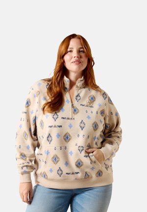 Woman with long red hair wearing beige pullover with blue and brown geometric patterns and light blue jeans, hand resting in front pocket.